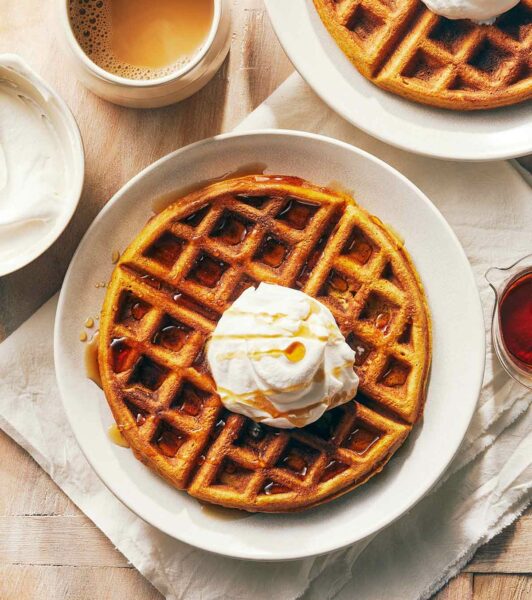 A wooden table with 2 plates of pumpkin waffles with maple whipped cream, alongside a bowl of whipped cream, maple syrup, and a cup of coffee.