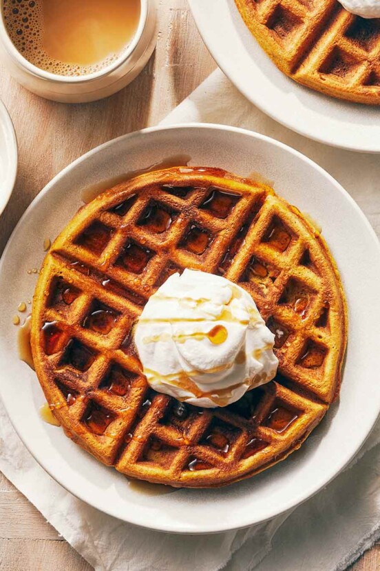 A wooden table with 2 plates of pumpkin waffles with maple whipped cream, alongside a bowl of whipped cream, maple syrup, and a cup of coffee.
