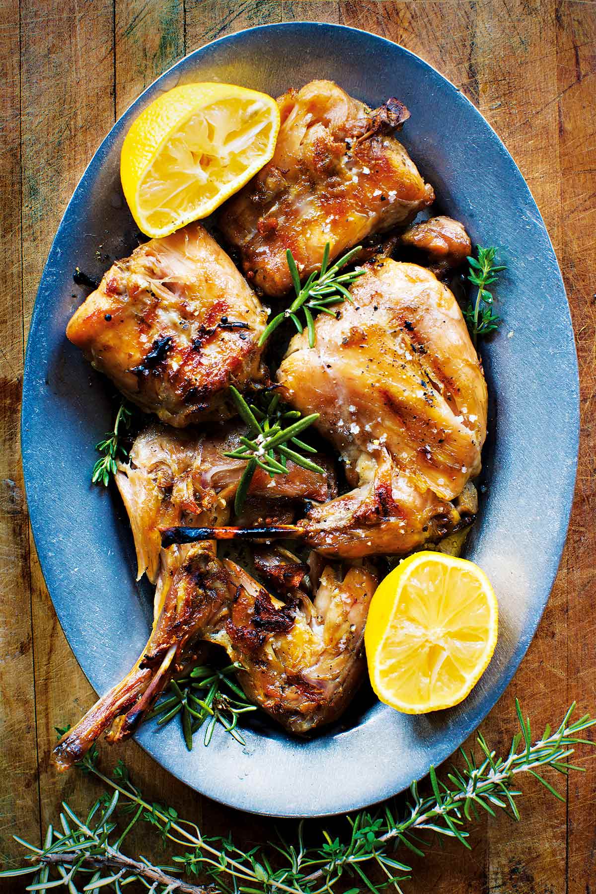 A blue platter filled with rabbit confit, garnished with twigs of rosemary and lemon halves.