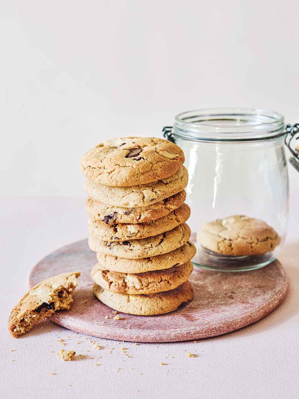 A wooden plate with a stack of salted tahini chocolate cookies, with a cookie jar and a partially eaten cookie.