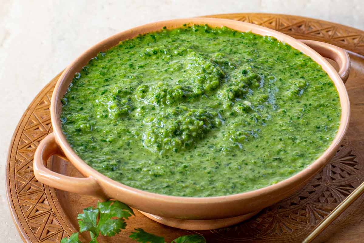 Schug in a pottery bowl, on a wooden plate with a spoon and garnished with cilantro.