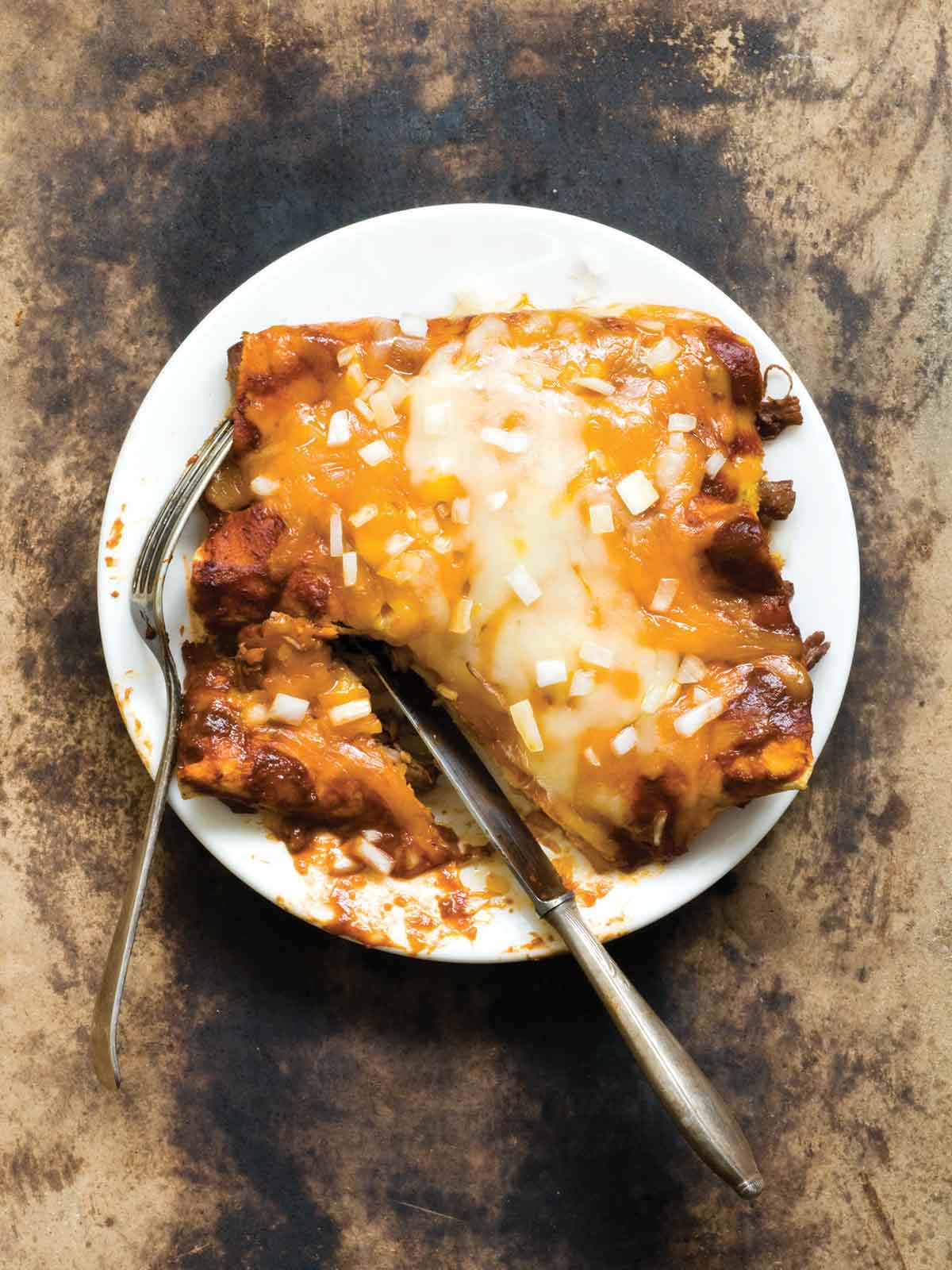 A white plate with 3 shredded beef enchiladas covered with melted cheese and diced onions, with a knife and fork.