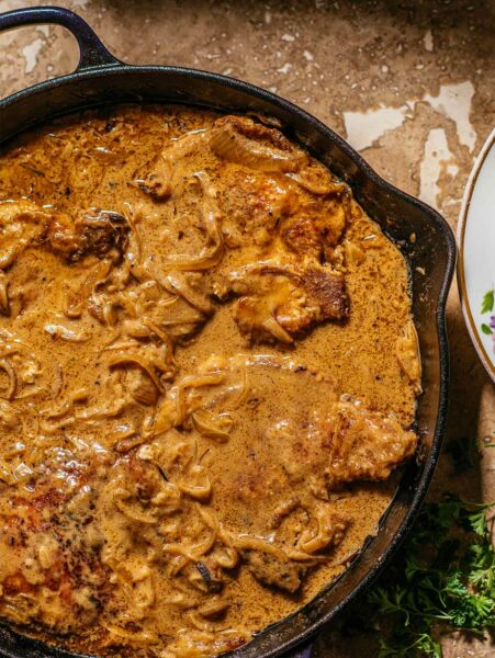 A cast-iron pan filled with smothered pork chops in a thick caramelized onion and cream sauce.