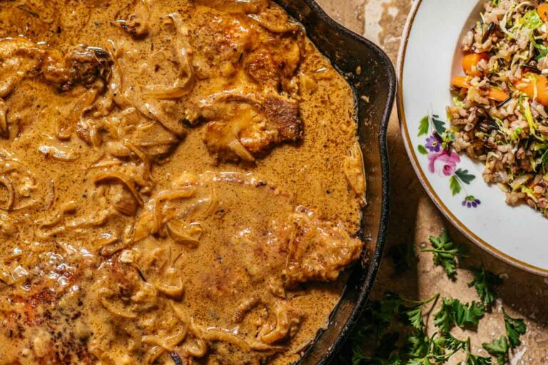 A cast-iron pan filled with smothered pork chops in a thick caramelized onion and cream sauce beside a plate of wild rice.