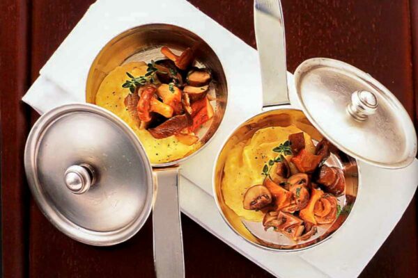 Soft polenta with forest mushrooms in two metal pots with lids.