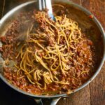 Spaghetti Bolognese in a large metal skillet, being served with tongs, on a wooden table.