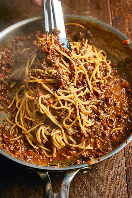 Spaghetti Bolognese in a large metal skillet, being served with tongs, on a wooden table.
