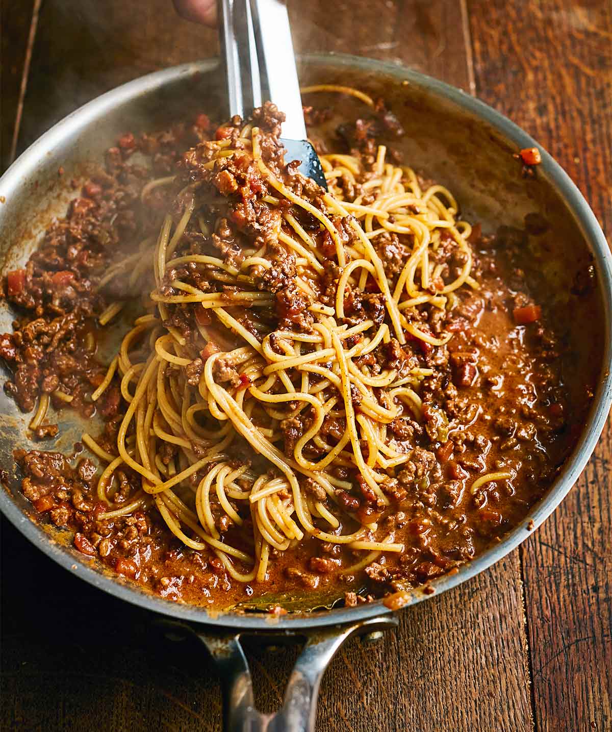 Spaghetti Bolognese in a large metal skillet, being served with tongs, on a wooden table.