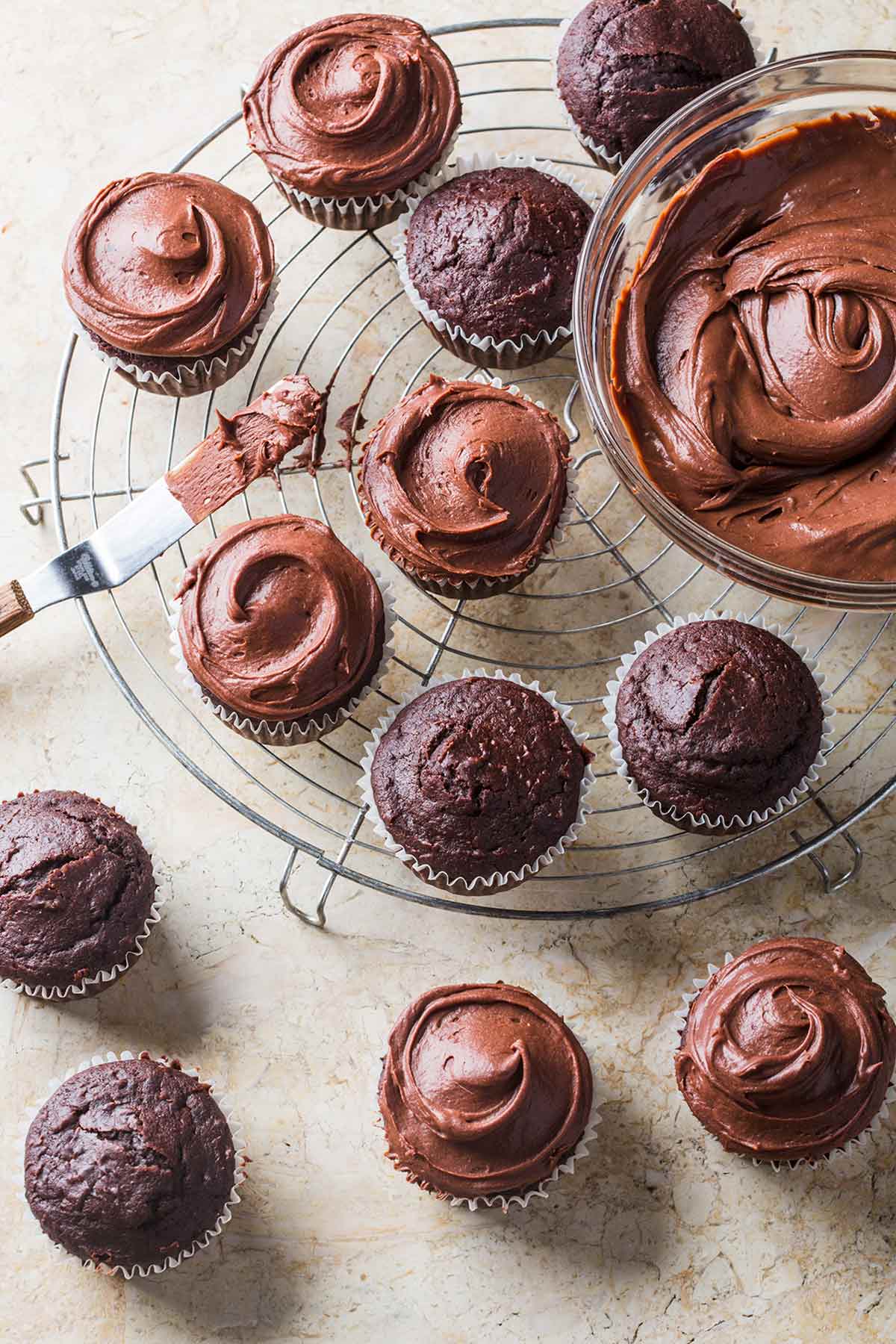 Vegan chocolate cupcakes, half frosted with vegan chocolate frosting, on a wire rack beside a bowl of frosting.