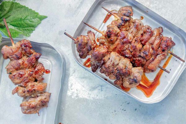 Two metal trays with skewers of Vietnamese pork skewers, garnished with Thai basil.