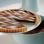 A white cake stand with a warm mocha tart on top, with cocoa and confectioner's sugar on top and one slice missing.