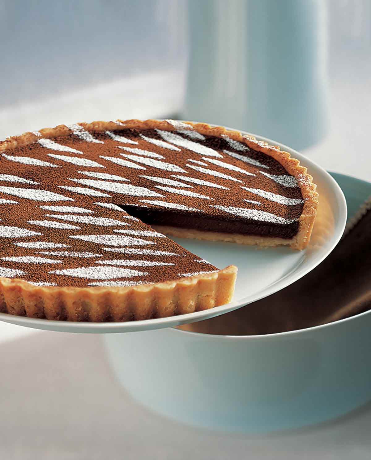 A white cake stand with a warm mocha tart on top, with cocoa and confectioner's sugar on top and one slice missing.