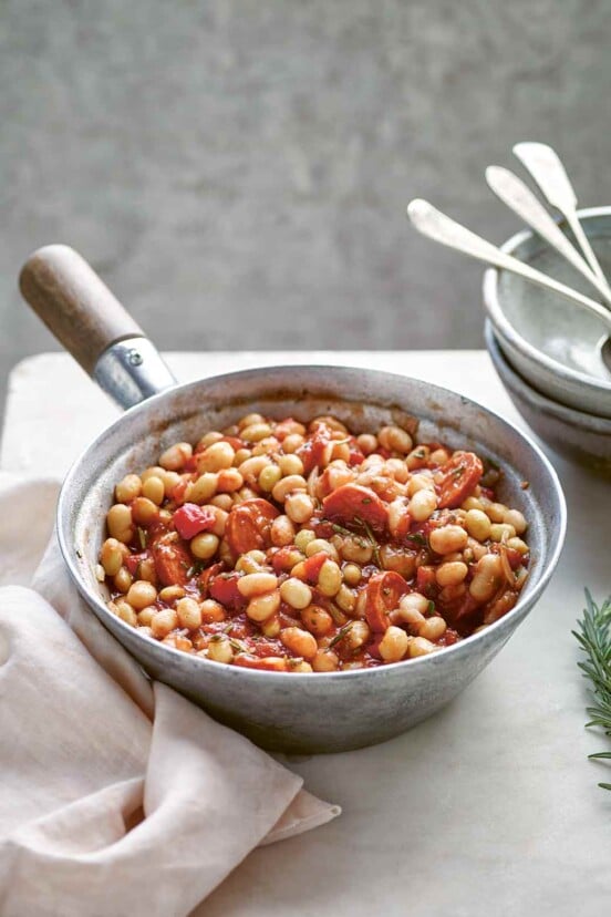 A metal saucepan filled with white bean stew with tomatoes and rosemary, with bowls, napkins, and utensils in the background.