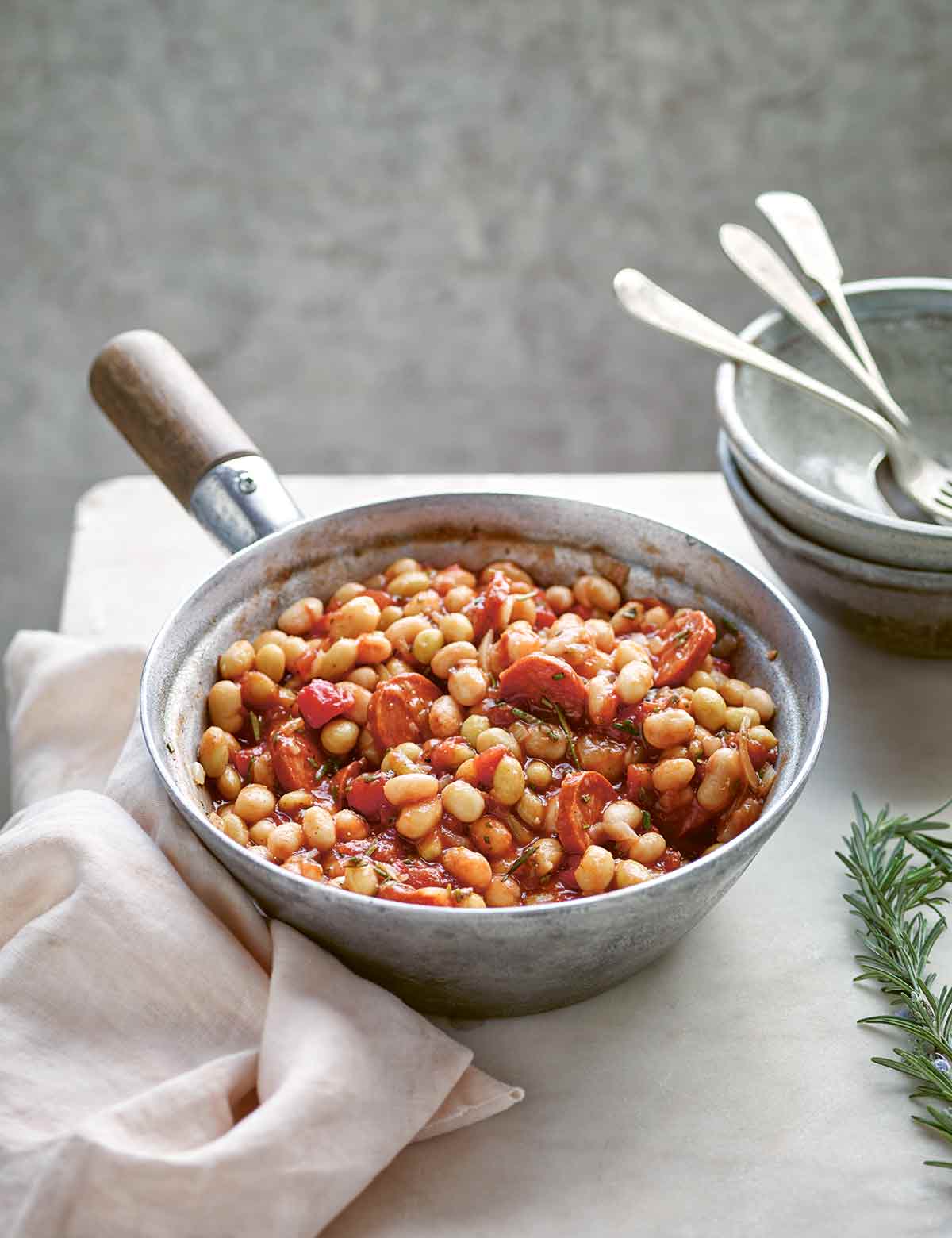 A metal saucepan filled with white bean stew with tomatoes and rosemary, with bowls, napkins, and utensils in the background.