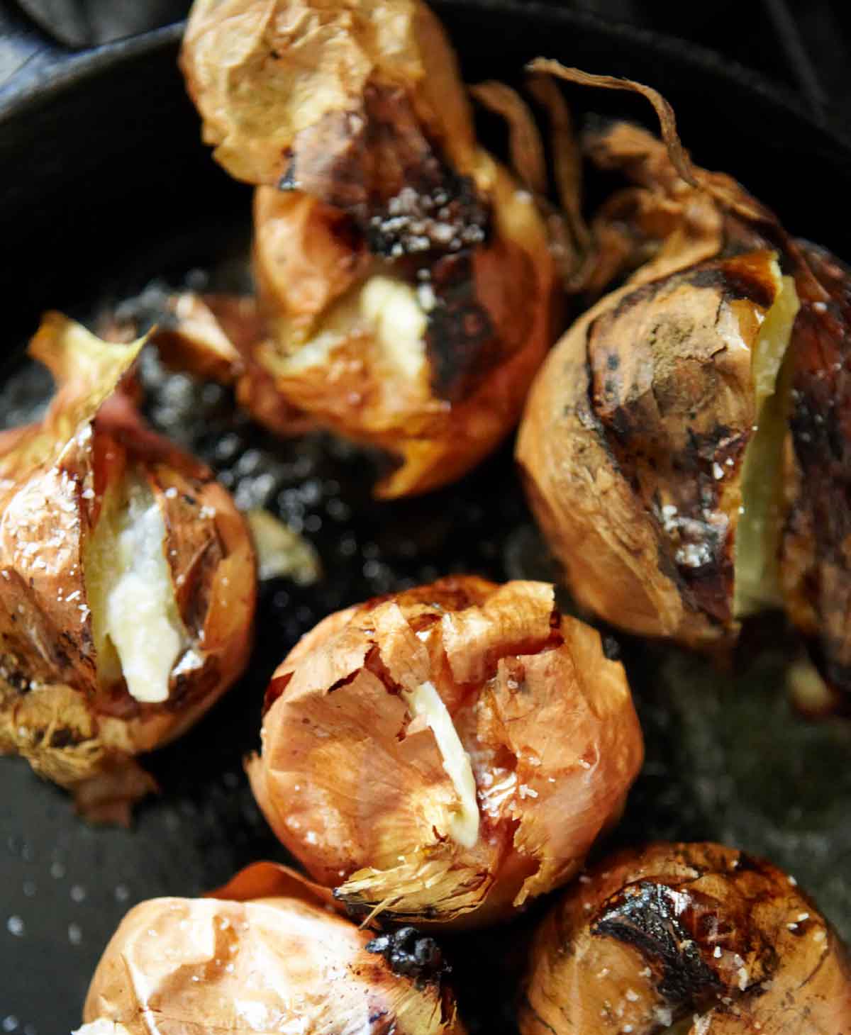 A cast-iron skillet with 8 whole roasted onions with papery skins and creamy interiors.