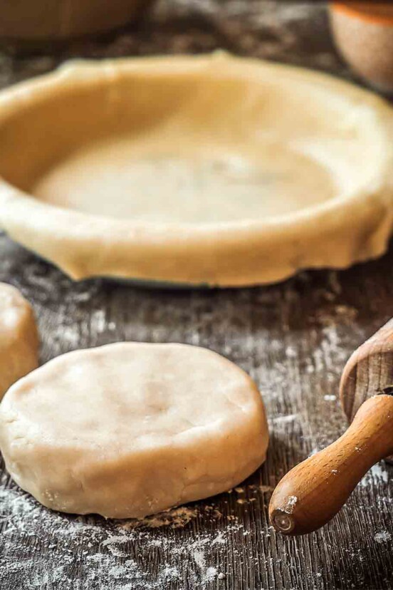 All butter pie dough in 2 rounds, with a pie plate and dough in the background, as well as a rolling pin.