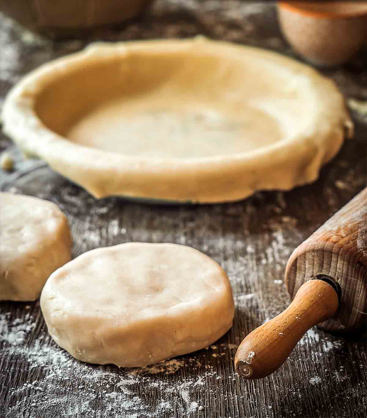 All butter pie dough in 2 rounds, with a pie plate and dough in the background, as well as a rolling pin.