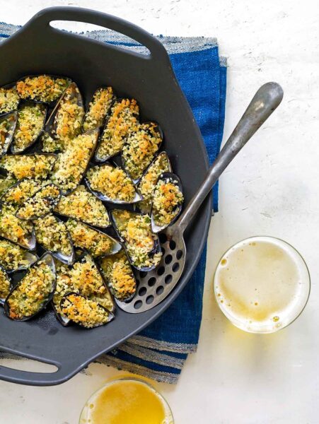 Baked mussels with breadcrumbs in a cast-iron pan with a serving spoon, on a blue tablecloth.