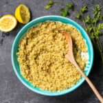 Baked quinoa with lemon and parsley in a turquoise bowl with a wooden spoon, surrounded by lemon halves and parsley sprigs.