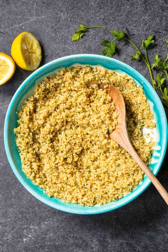 Baked quinoa with lemon and parsley in a turquoise bowl with a wooden spoon, surrounded by lemon halves and parsley sprigs.