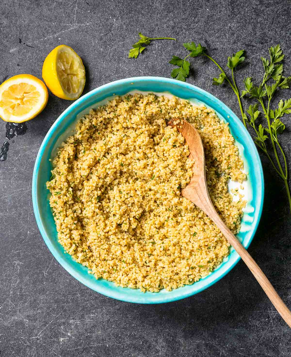 Baked quinoa with lemon and parsley in a turquoise bowl with a wooden spoon, surrounded by lemon halves and parsley sprigs.