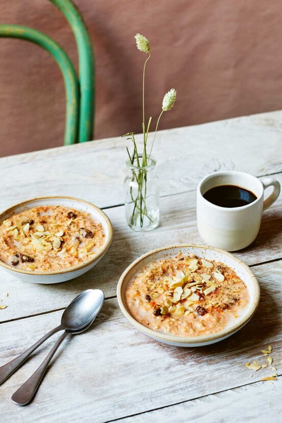 Carrot cake oatmeal in two bowls on a wooden table, beside a cup of black coffee, two spoons, and a vase of flowers.