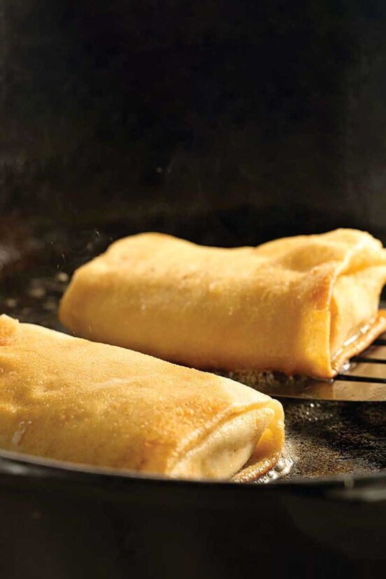 Cheese blintzes in a cast-iron frying pan, one being lifted up by a metal spatula.