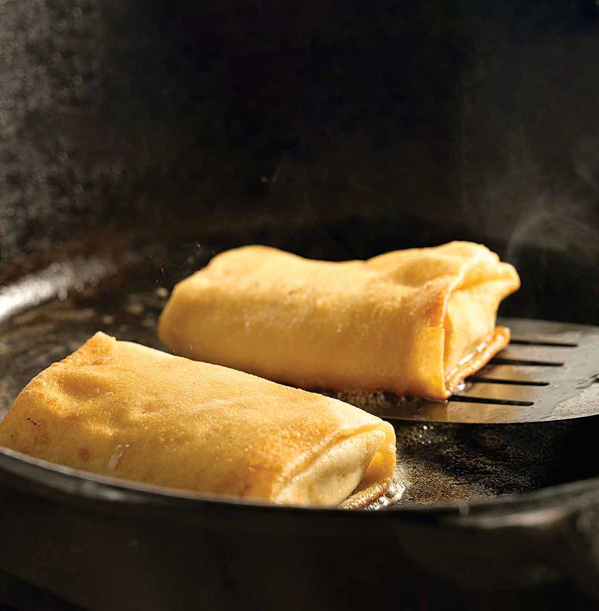 Cheese blintzes in a cast-iron frying pan, one being lifted up by a metal spatula.