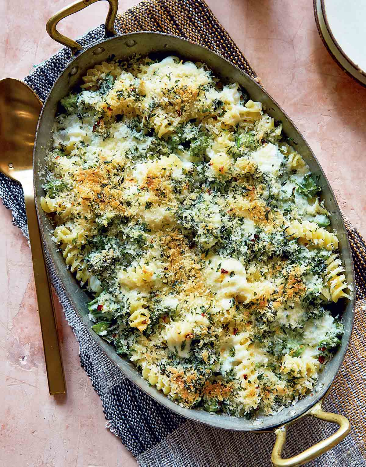 Baked cheesy pasta with broccoli in a large metal casserole dish, with a serving spoon and napkin.