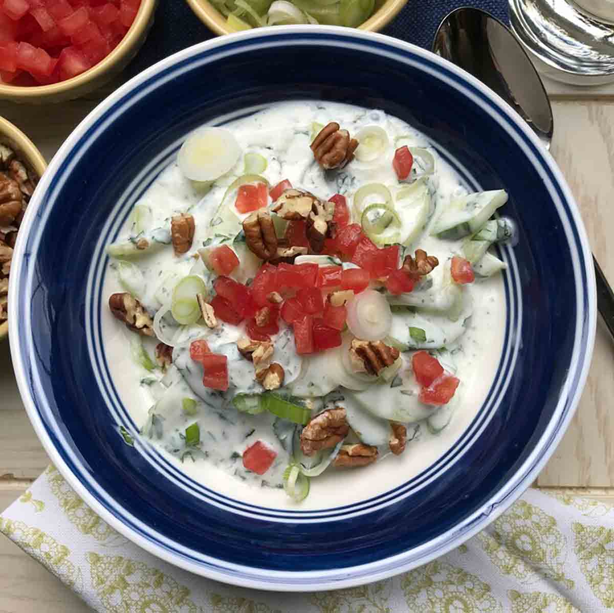 Chilled cucumber soup in a blue and white bowl, topped with red peppers, green onions, and chopped pecans.
