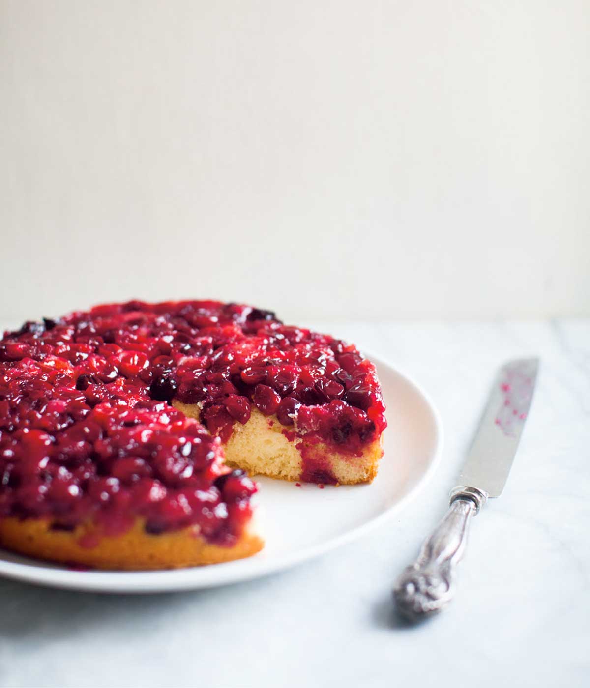 Cranberry upside-down cake with a large piece missing, on a white plate with a serving knife beside it.