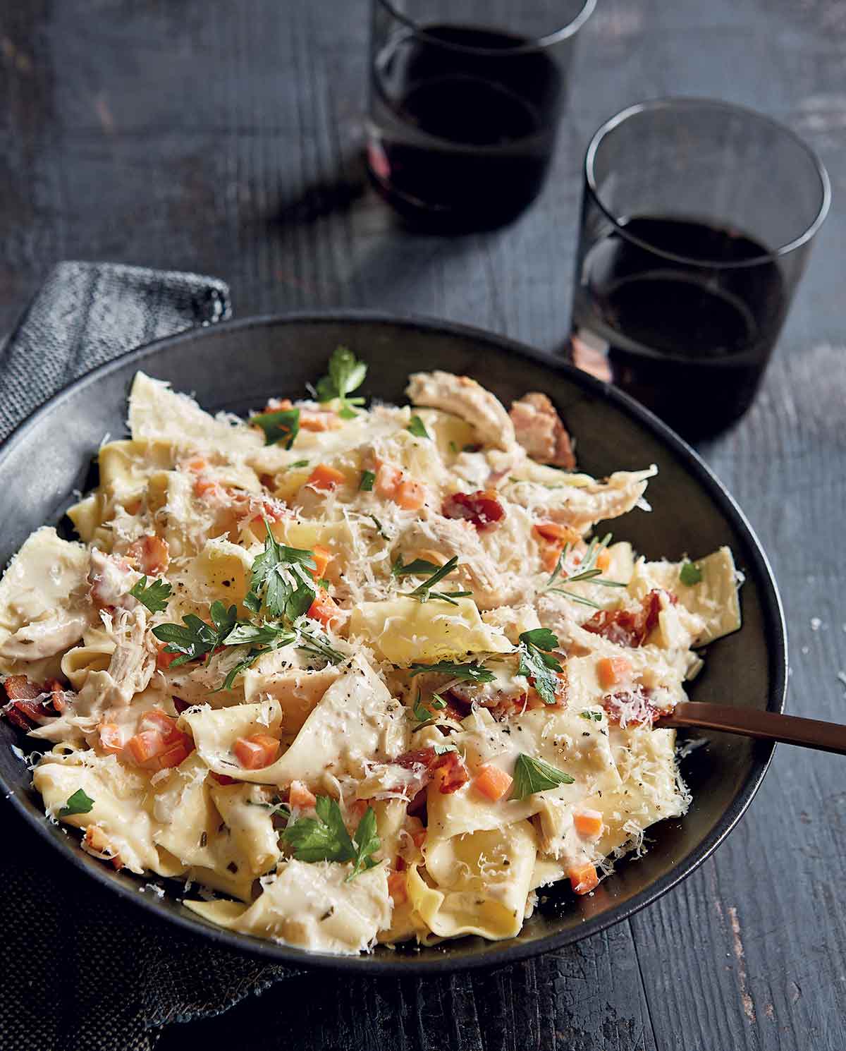 Creamy pappardelle with chicken and bacon in a black bowl with a fork, topped with grated cheese.