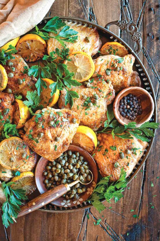 Crispy chicken with lemon and capers in a large serving tray, with a bowl of capers and a bowl of peppercorns. The chicken is garnished with lemon slices and parsley.