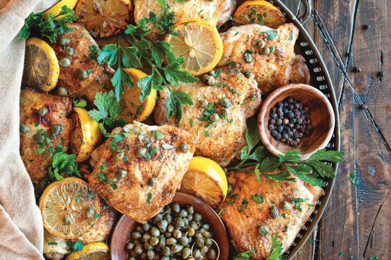Crispy chicken with lemon and capers in a large serving tray, with a bowl of capers and a bowl of peppercorns. The chicken is garnished with lemon slices and parsley.
