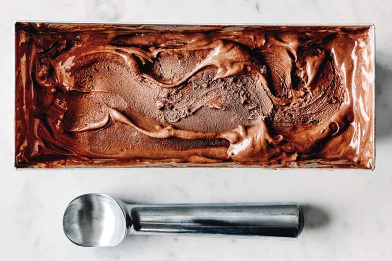 Dark chocolate and tahini ice cream in a metal ice cream tin with a swirl on top, with a metal ice cream scoop laying beside it.