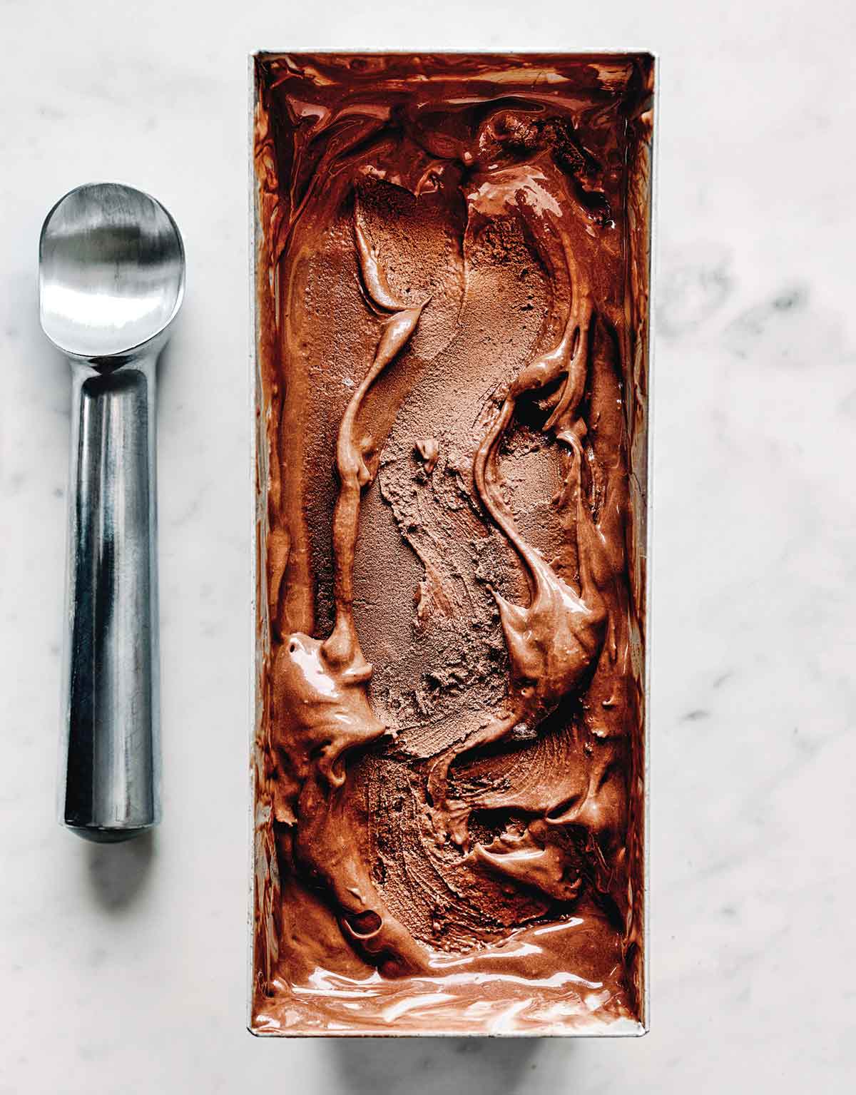 Dark chocolate and tahini ice cream in a metal ice cream tin with a swirl on top, with a metal ice cream scoop laying beside it.