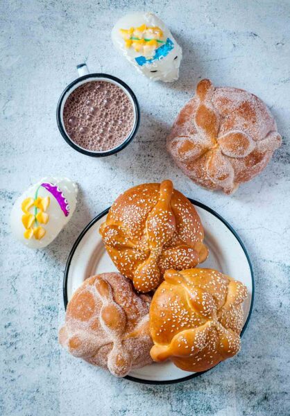 Day of the Dead bread on a plate, 2 with sugar and 2 with white sesame seeds, beside a cup of Mexican hot chocolate and 2 sugar skulls.