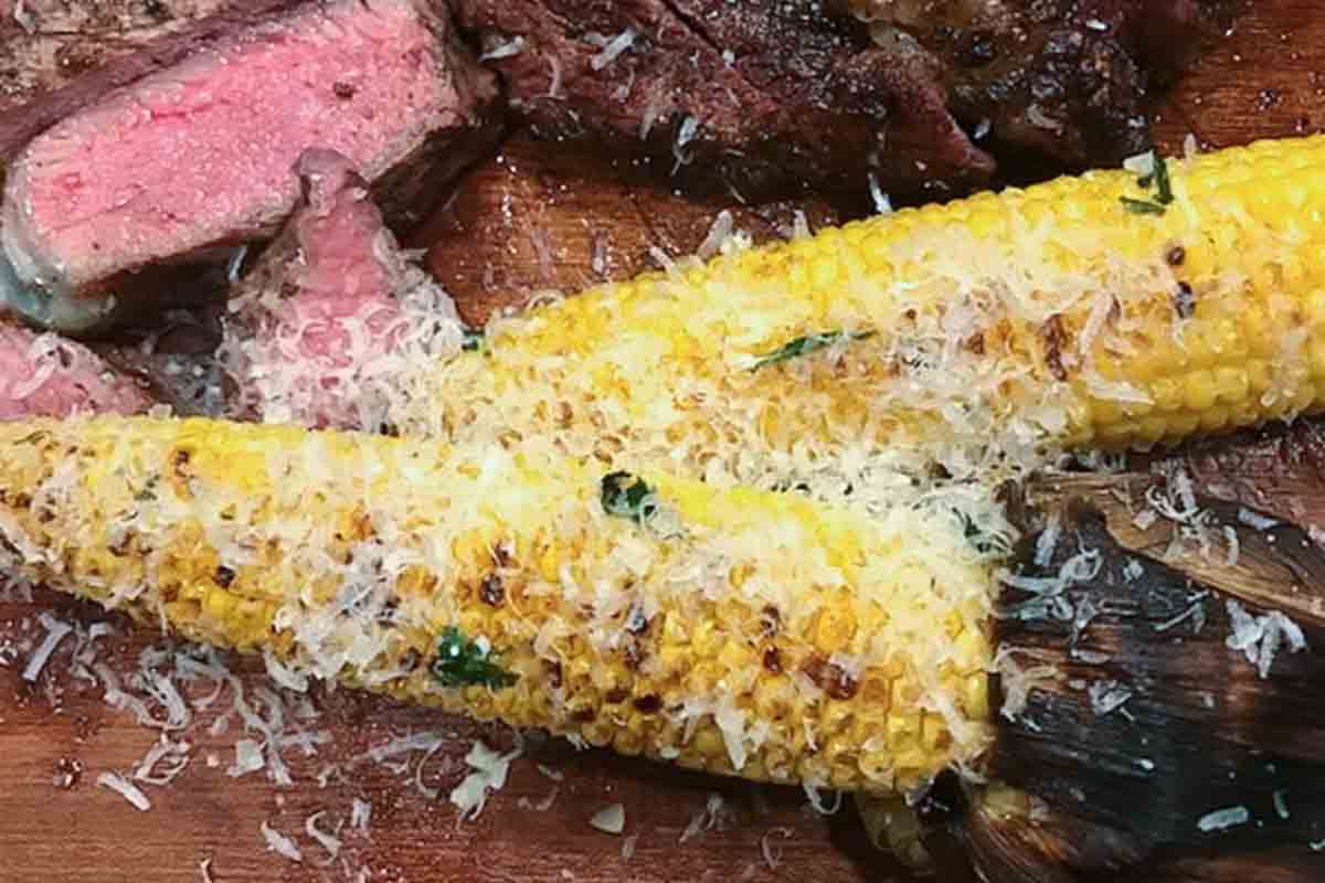 Elote garnished with grated cheese on a cutting board with slices of rare steak.