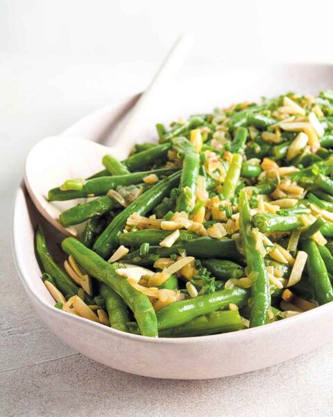 Green beans with browned butter and almonds in an oval white bowl with a serving spoon.