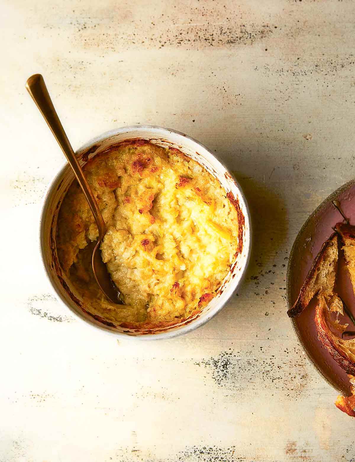 Hot artichoke dip in a white pottery bowl with a serving spoon, beside a bowl of toasted bread.