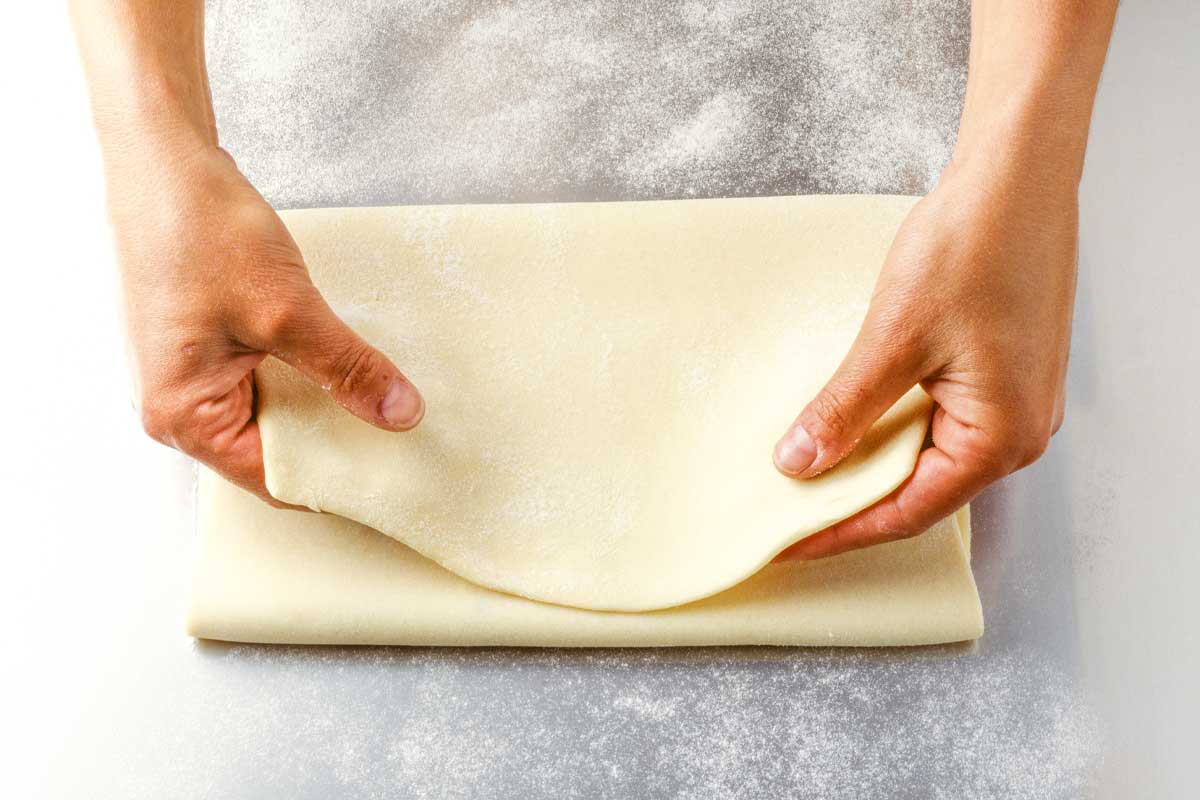 Extra flaky pie crust being folded in thirds from a large sheet of dough on a floured surface.