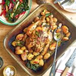 Lemon and herb roast chicken with fingerling potatoes and cloves of garlic, in a roasting pan, beside a plate of Swiss chard salad.