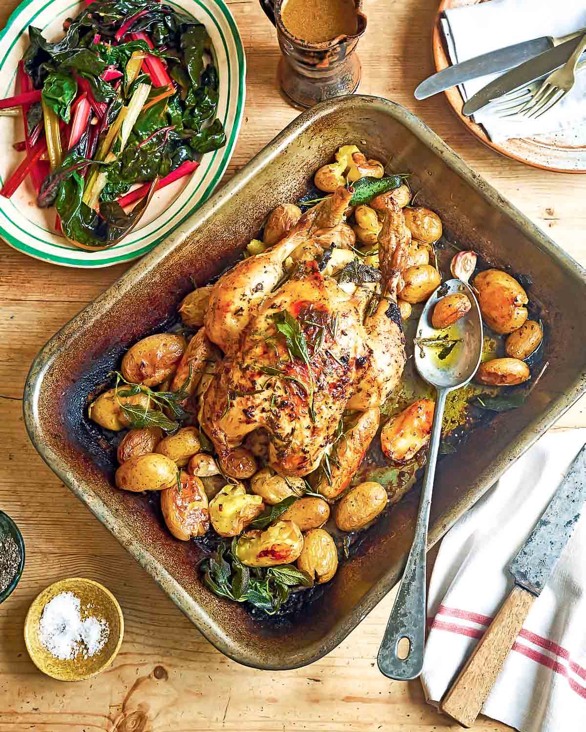 Lemon herb roast chicken with fingerling potatoes and cloves of garlic, in a roasting pan, beside a plate of Swiss chard salad.