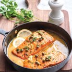 Lemon salmon piccata, 2 pieces, in a metal pan with slices of lemon, pan sauce, garnished with garlic and capers.