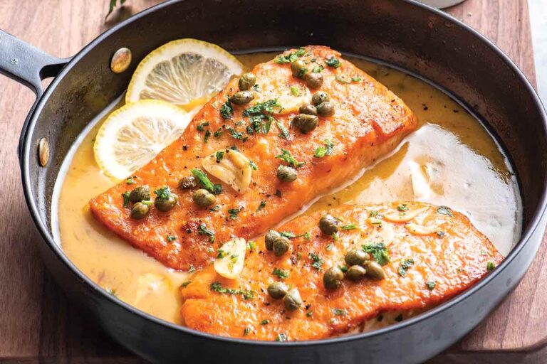 Lemon salmon piccata, 2 pieces, in a metal pan with slices of lemon, pan sauce, garnished with garlic and capers.