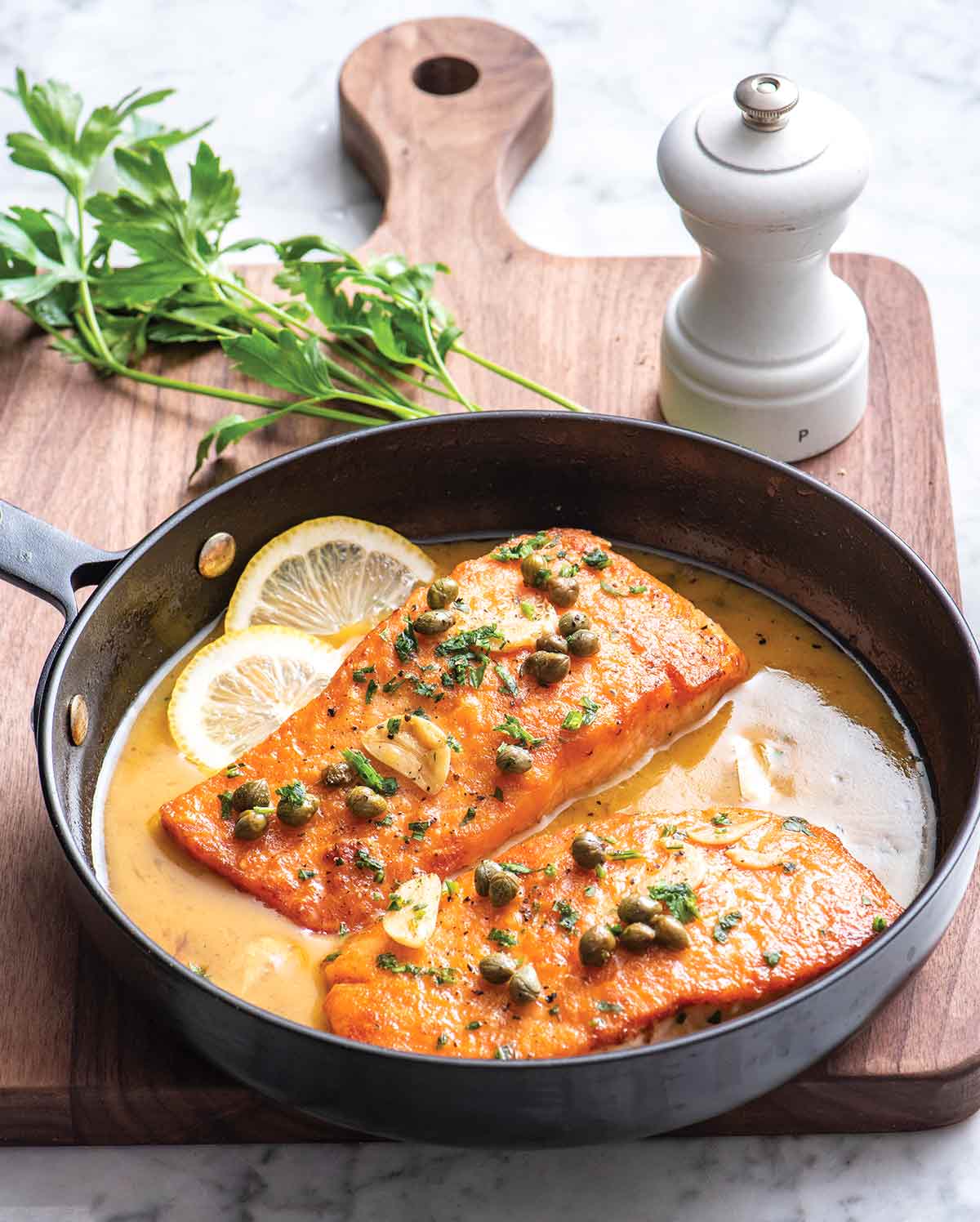 Lemon salmon piccata, 2 pieces, in a metal pan with slices of lemon, pan sauce, garnished with garlic and capers.