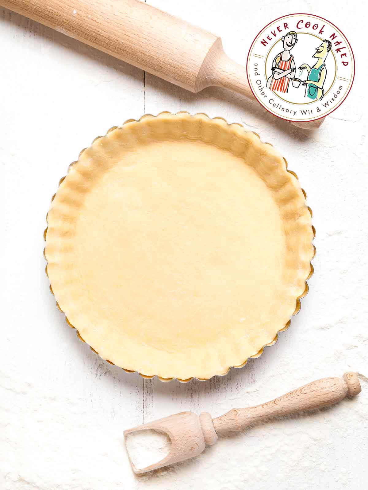 How to stop pastry from shrinking, a pie crust in a fluted pie tine beside a flour scoop and a rolling pin.