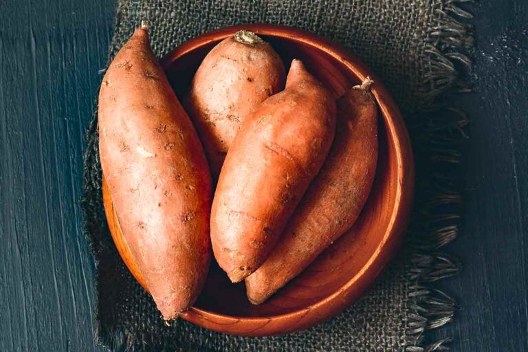 Four sweet potatoes in a wooden bowl on a piece of burlap.