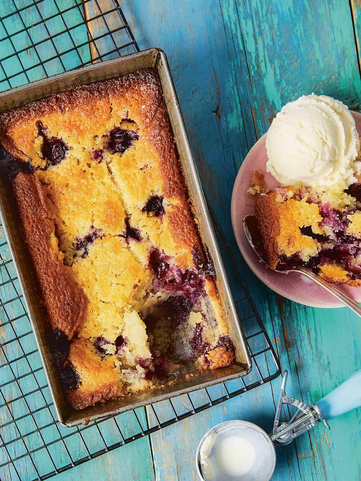 One-bowl blueberry buckle in a metal loaf pan cooling on a wire rack, with a pink bowl filled with buckle and a scoop of ice cream.