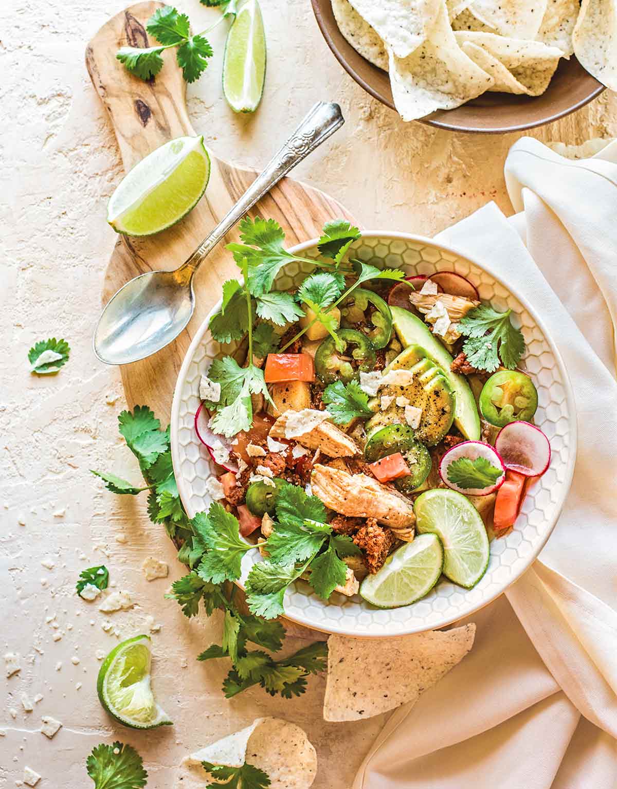 Paleokyckling- och chorizogryta i en skål, garnerad med lime, avokado, koriander och rädisor, bredvid en skål med tortillachips.